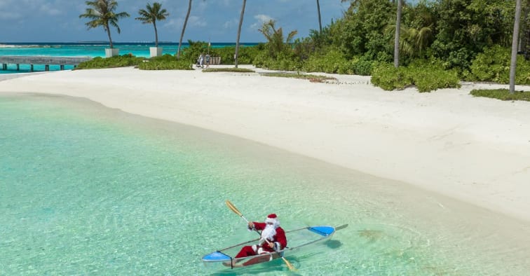 Santa Claus kayaking in clear turquoise waters near a white sand beach at Niyama Private Islands Maldives.